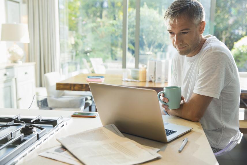 A man does reads from a laptop while drinking his morning coffee in his kitchen.